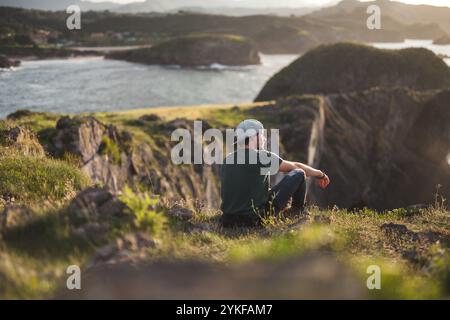 Un jeune homme est assis paisiblement au sommet d'une falaise accidentée des Asturies, contemplant la côte pittoresque baignée de soleil doré Banque D'Images