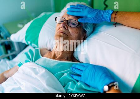Une femme âgée allongée dans un lit d'hôpital reçoit une touche de compassion d'un professionnel de la santé portant des gants bleus le cadre met l'accent sur Medi Banque D'Images