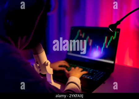 Vue arrière d'un garçon méconnaissable engagé dans le commerce en ligne. Il est assis dans une pièce sombre avec un éclairage coloré, se concentrant sur les graphiques boursiers Banque D'Images