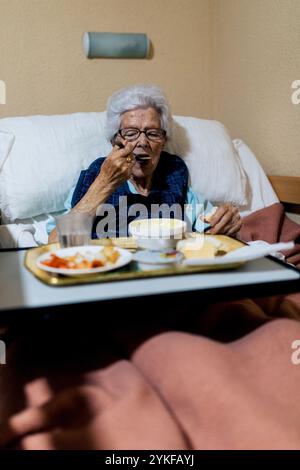 Une femme âgée profite d'un repas dans sa confortable salle de soins infirmiers, assis dans le lit avec un plateau le cadre chaleureux et accueillant met en évidence le quotidien Banque D'Images