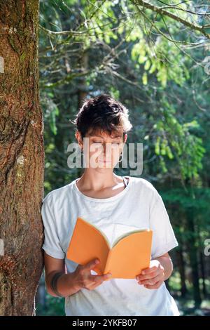 Un individu non binaire profondément engagé dans la lecture d'un livre orange, appuyé contre un arbre dans une forêt luxuriante, avec la lumière du soleil filtrant à travers les feuilles. Banque D'Images