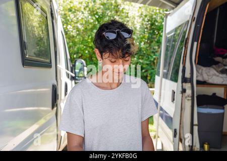 Une personne non binaire dans une tenue décontractée et des lunettes de soleil poussées sur ses cheveux se tient à côté d'une camionnette, regardant vers le bas avec une expression douce, au milieu d'un dos Banque D'Images