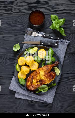 la moitié de poulet rôti avec des pièces d'épis de maïs et des feuilles de basilic frais sur l'assiette sur une table en bois noir avec couverts et trempage de chili doux, vue verticale Banque D'Images