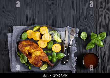 la moitié du poulet grillé avec des pièces d'épis de maïs et des feuilles de basilic frais sur l'assiette sur une table en bois noir avec couverts et sauce chili douce, vi horizontale Banque D'Images