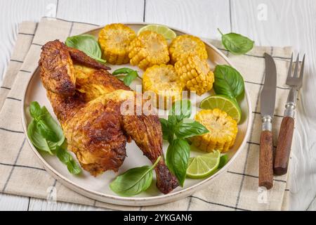 la moitié du poulet rôti avec des pièces d'épis de maïs et des feuilles de basilic frais sur l'assiette sur une table en bois blanc avec des couverts, vue horizontale, gros plan Banque D'Images