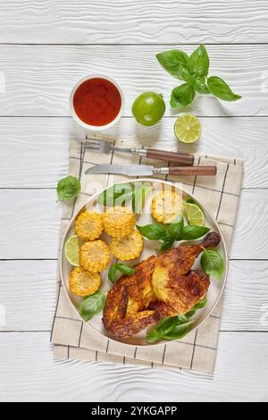 la moitié du poulet bbq avec des pièces d'épis de maïs et des feuilles de basilic frais sur l'assiette sur une table en bois blanc avec couverts, citron vert et sauce chili doux, vue verticale Banque D'Images