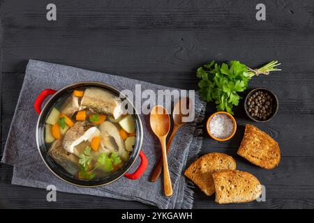 Soupe claire de brochet avec légumes, persil et épices dans un pot rouge sur une table en bois noir avec cuillères en bois et pain grillé, vue horizontale d'en haut, Banque D'Images