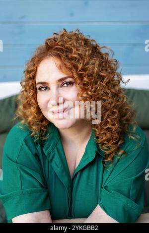 Femme de taille plus avec cheveux rouges bouclés en vêtements verts contre fond de mur en bois bleu coloré, beauté et confiance en extérieur Banque D'Images