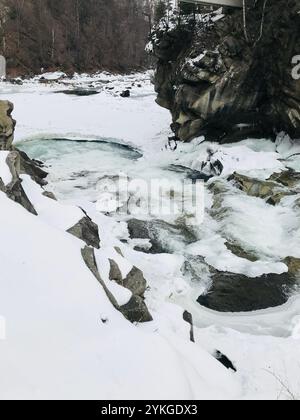 Une belle scène hivernale avec une rivière gelée serpentant à travers des rochers chargés de neige, créant une atmosphère tranquille et sereine dans une région boisée. Banque D'Images