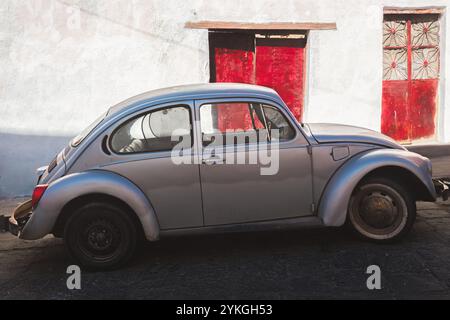Guanajuato, Mexique - 6 février 2024 : une Volkswagen Beetle argentée vintage avec une patine altérée garée à côté d'un bâtiment rustique avec DO rouge frappant Banque D'Images