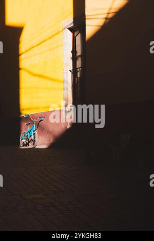 Un vélo bleu solitaire repose contre un mur endommagé dans une ruelle calme et ensoleillée de la ville historique de Guanajuato, projetant de profondes ombres qui soulignent les contras Banque D'Images