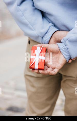 Gros plan de la main d'un jeune homme tenant une petite boîte rouge dans son dos. Jeune homme tenant un cadeau de saint Valentin pour sa petite amie Banque D'Images