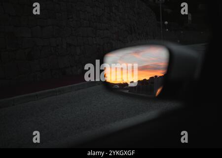 Rétroviseur latéral d'une voiture capturant les couleurs vives d'un coucher de soleil, avec un mur de pierre et une route en arrière-plan Banque D'Images