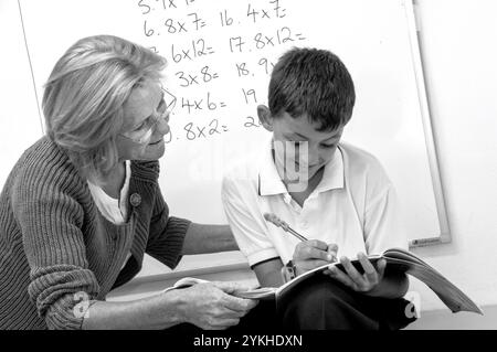 Professeur de maths et garçon de l'école primaire 7-10 ans et tableau blanc travaillant sur les tables de somme mathématiques fois avec l'aide d'une enseignante dans la salle de classe de l'école. Tableau blanc de classe d'école tables de multiplication des temps. (Traitement noir et blanc Alt) Banque D'Images