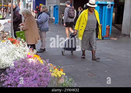 Chariot de magasinage senior plus âgé femme âgé portant chapeau de paille manteau jaune marchant dans la rue par l'étal de fleurs à Brixton Londres Angleterre KATHY DEWITT Banque D'Images