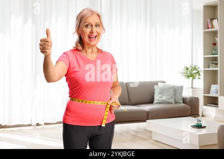 Heureuse femme mature mesurant sa taille et montrant les pouces vers le haut dans un salon Banque D'Images