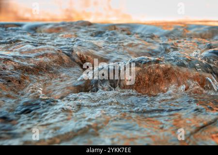 L'eau se précipite sur des roches lisses, capturant le mouvement dynamique d'un ruisseau qui coule. La lumière du soleil se reflète sur l'eau, lui donnant une lueur chaude et vibrante. Banque D'Images