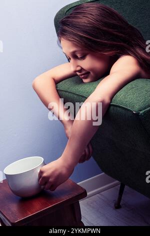 Jeune fille se relaxant paisiblement sur un fauteuil confortable, ressemblant à un chat alors qu'elle tend pour une tasse sur une table dans son intérieur serein. Elle incarne le chil Banque D'Images