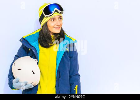 Concept d'activité de sport d'hiver. Belle femme souriante portant une veste bleue, des lunettes de ski et un pull vert tenant le casque, fond bleu studio shot. Banque D'Images