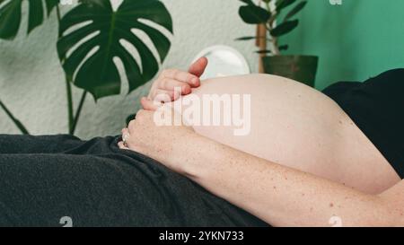 La future Mother place doucement son bébé Bump à la maison dans un cadre confortable avec une végétation luxuriante Banque D'Images