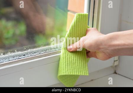 La main de la femme essuie la condensation de l'eau sur la fenêtre blanche dans la salle de bain avec un niveau d'humidité élevé. Banque D'Images