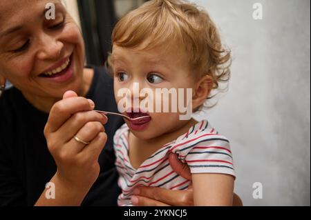 Une mère nourrit avec amour son fils tout-petit, qui a l'air curieux tout en dégustant de la nourriture à la cuillère. Le cadre intérieur confortable ajoute de la chaleur à l'instant présent, highlig Banque D'Images