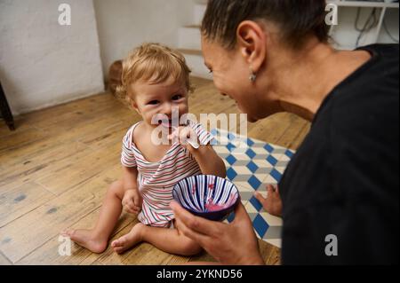 Une scène délicieuse d'un parent nourrissant joyeusement leur tout-petit dans un cadre confortable à la maison. Le sourire éclatant de l'enfant et le bol coloré de nourriture créent un Banque D'Images