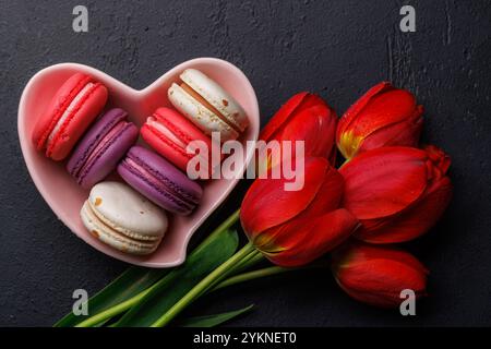 Tulipes rouges et macarons vibrants remplissent un bol en forme de cœur, offrant une scène charmante et charmante parfaite pour les célébrations ou les moments joyeux Banque D'Images