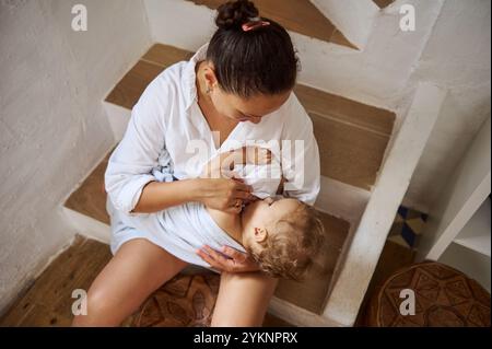 Un moment tendre où une mère allaite son bébé assis dans les escaliers. La scène capture la chaleur, l'éducation, et le lien maternel dans une maison confortable env Banque D'Images