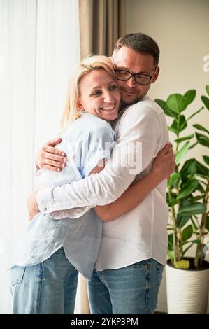 Couple heureux embrassant sur fond clair à la maison. Jolie femme souriant et embrassant son mari. Couple doux en vêtements décontractés. Banque D'Images