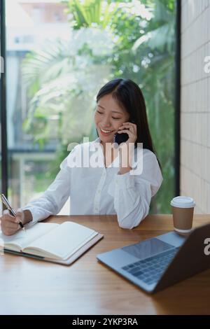Une jeune femme sourit en parlant au téléphone, notant des notes dans son carnet. Un ordinateur portable élégant et une tasse à café reposent sur la table en bois, créant Banque D'Images