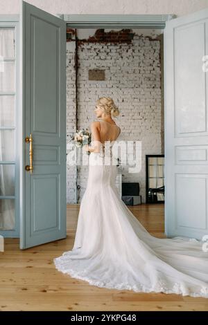 Une belle mariée dans une robe élégante tient des fleurs vibrantes, debout par des portes ouvertes menant à une célébration joyeuse Banque D'Images