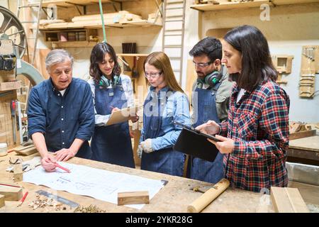 Un groupe diversifié de travailleurs dans un chantier forestier, faisant preuve de travail d'équipe et de formation. Les jeunes apprentis apprennent de mentors expérimentés autour d'un atelier c Banque D'Images