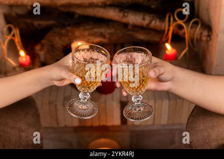 Une scène de soirée confortable avec deux mains levant des verres de cristal dans un toast près de la cheminée, entouré par la lueur chaude des bougies. Banque D'Images