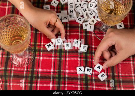 Deux mains arrangent les carreaux de lettres orthographiant « Joyeux Noël » sur une nappe à carreaux rouges, entourés de carreaux éparpillés et de verres à vin. Banque D'Images