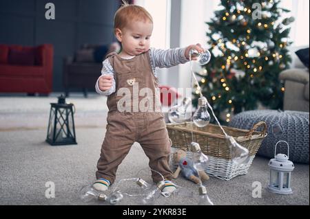 Jeune enfant joue avec des ampoules près d'un arbre de Noël décoré dans un cadre de salon confortable pendant l'hiver Banque D'Images