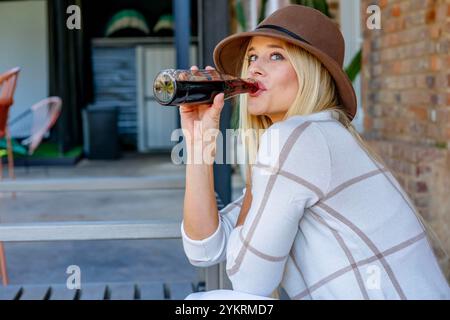 Une superbe jeune blonde dans une tenue chic et décontractée profite d'un soda dans un centre commercial en plein air. Le soleil la baigne dans la chaleur comme elle prend dans l'énergie vibrante et s. Banque D'Images