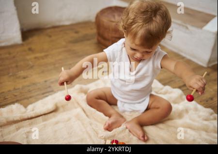 Un tout-petit mignon dans une tenue blanche joue joyeusement avec un jouet rouge sur un plancher en bois, mettant en valeur l'exploration de l'enfance. Banque D'Images