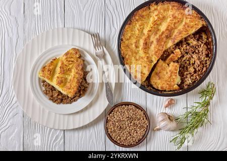 tarte végétalienne aux bergers de lentilles cuites surmontée de purée de pommes de terre sur une assiette blanche avec fourchette et dans un plat de cuisson sur une table en bois, vue horizontale de dessus Banque D'Images