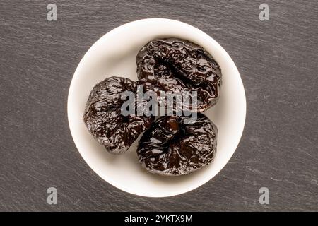 Pruneaux sucrés dans une assiette en céramique sur une pierre d'ardoise, macro, vue de dessus. Banque D'Images