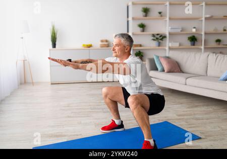 Restez seul à la maison pour vous entraîner. Fit homme mûr dans des vêtements de sport faisant des squats dans l'intérieur du salon, exerçant ses jambes Banque D'Images