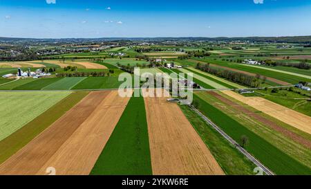 Vue aérienne de vastes terres agricoles avec des parcelles de champs verts, bruns et or sous un ciel bleu clair. Les bâtiments agricoles parsèment le paysage, illustrant le charme agricole et la diversité de la région. Banque D'Images