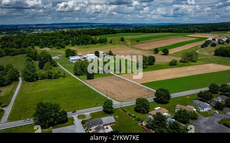 De vastes champs s'étendent à travers l'horizon, avec des parcelles de cultures brunes et vertes sous un ciel partiellement nuageux. Une route voisine longe les terres agricoles, reliant une zone résidentielle. Banque D'Images