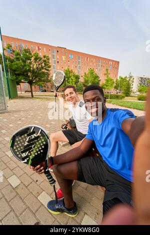 Deux jeunes hommes sont assis sur un banc, l'un d'eux prenant un selfie avec un téléphone portable. Banque D'Images