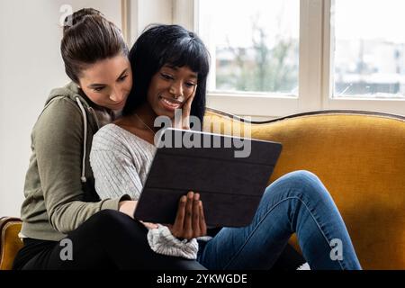 Couple LGBTQ+ aimant profiter d'un moment confortable ensemble tout en regardant une tablette. Deux femmes souriantes et souriantes, partageant du contenu sur un appareil numérique dans Banque D'Images