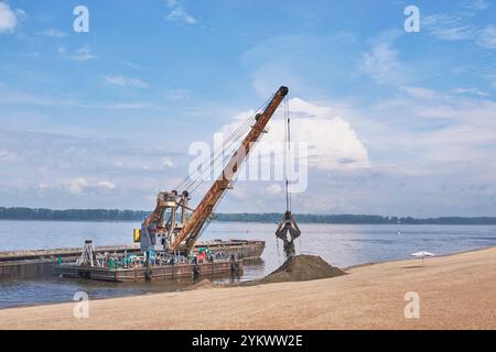 Samara, Russie - 8 juin 2024 : grue flottante sur la rivière calme Volga décharge le sable de la barge à la plage de sable par temps clair. Contrastes de machines industrielles Banque D'Images