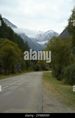 Une route alpine tranquille serpentant à travers une vallée luxuriante, encadrée par des arbres verts et menant vers de majestueuses montagnes enneigées sous un ciel couvert Banque D'Images