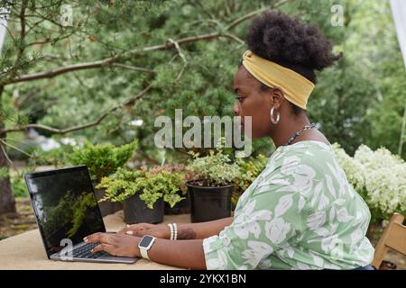 Portrait de vue de côté de la jeune femme afro-américaine à l'aide d'un ordinateur portable dans la plantation verte et dactylographiant tout en gérant la petite entreprise à thème écologique Banque D'Images