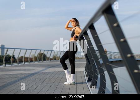 Une jeune femme s'exerçant sur une promenade élégante sous un ciel clair, elle protège ses yeux du soleil, incarnant un mélange de concentration et d'élégance dans son activité Banque D'Images