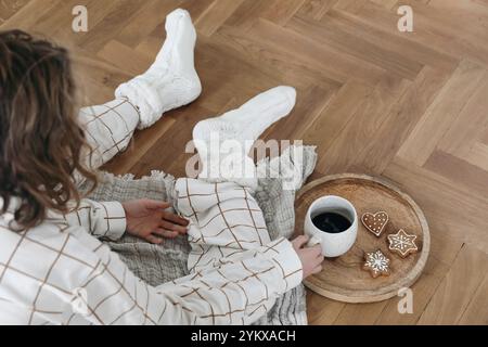 Natures mortes de Noël. Enfant en pyjama à carreaux assis sur du parquet. Jambes en chaussettes en laine. Tasse de café, biscuits au pain d'épices sur plateau. Chêne flou Banque D'Images